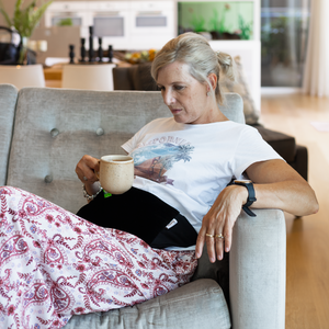 older woman relaxing on couch drinking tea wearing the hottle heat pack on stomach
